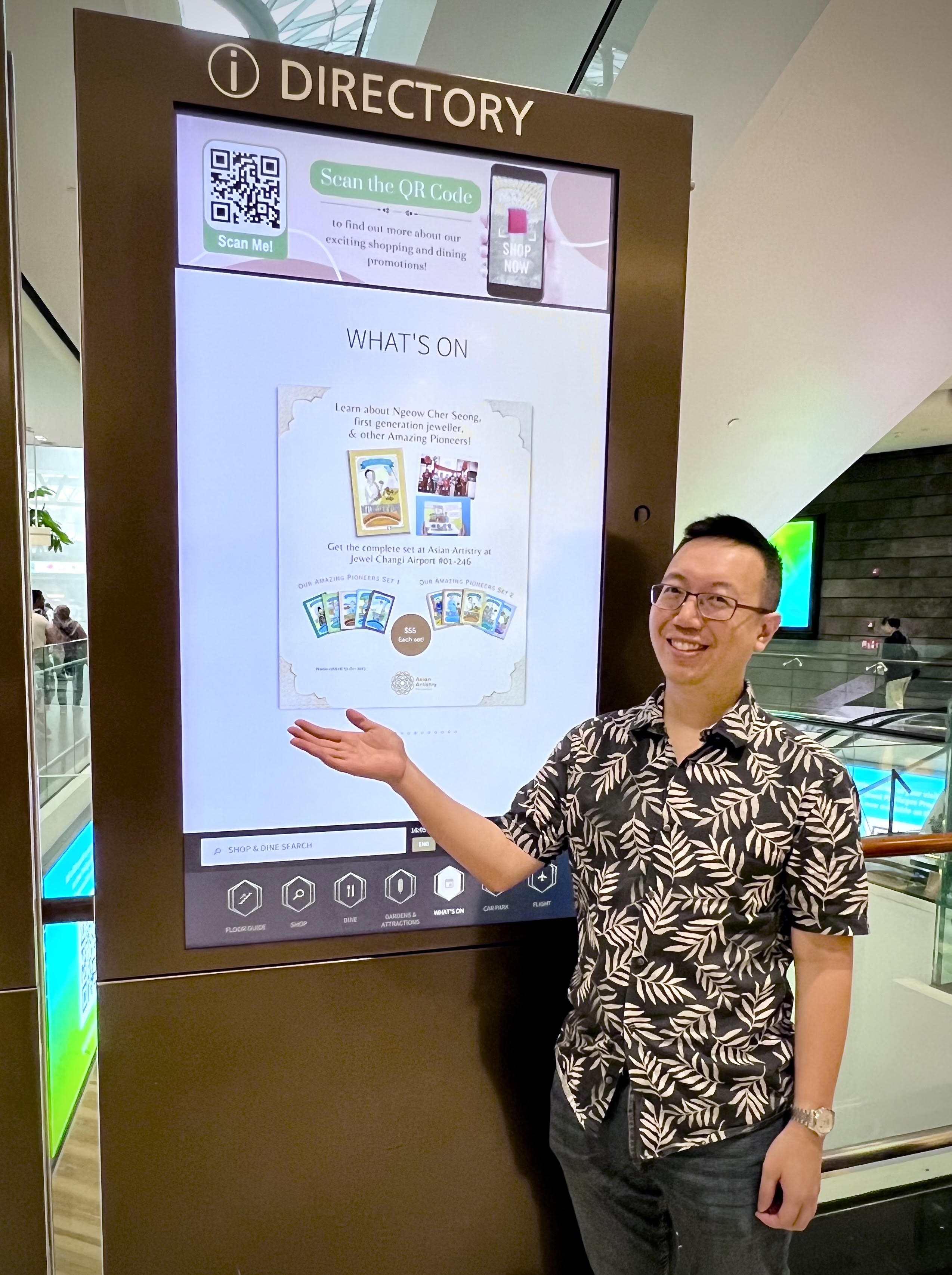 Shawn Seah smiling as he stands next to a digital board at Jewel, Changi Airport, that shows an advertisement covering his books, Our Amazing Pioneers. 