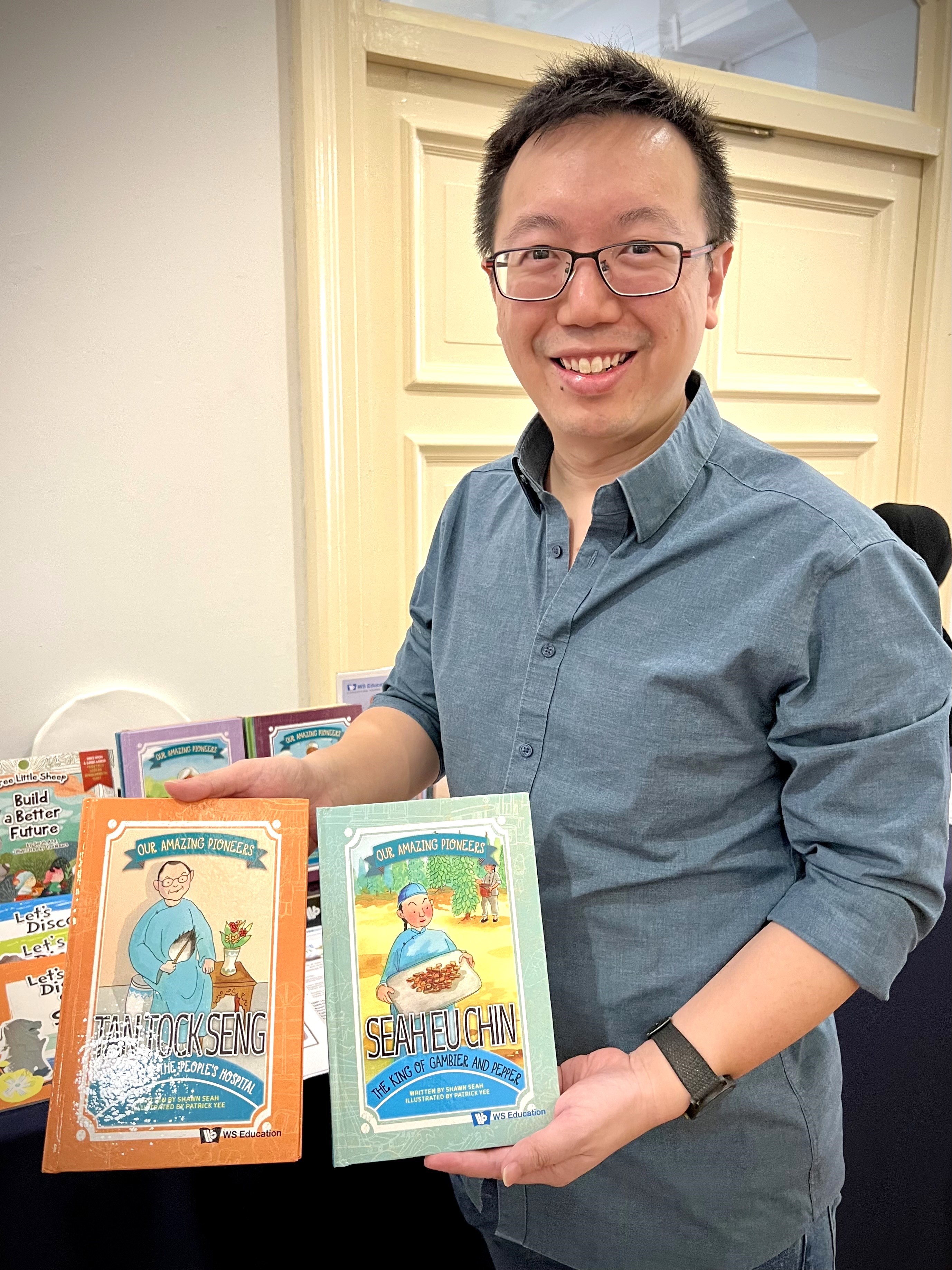 Published author Shawn Seah standing in front of a book booth, holding up Our Amazing Pioneers.